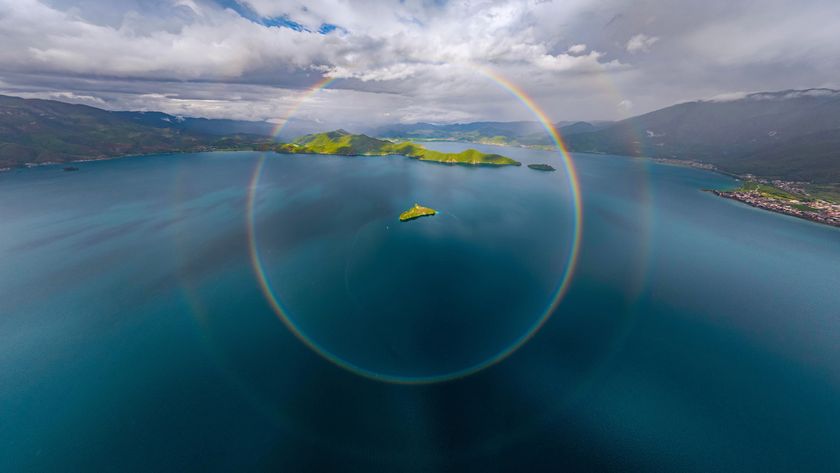 A stunning aerial view of a serene lake surrounded by green hills, featuring a vibrant rainbow halo overhead