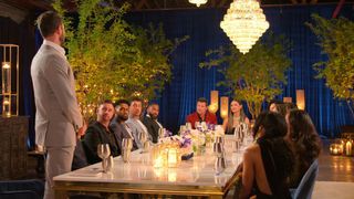 A man stands and addresses a table of 14 people, with men on the left, women on the right, and hosts Nick and Vanessa Lachey at the head, on 'The Ultimatum' season 3.