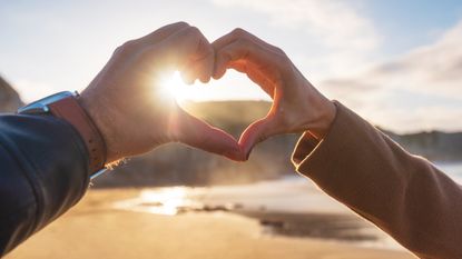 A couple forms the shape of a heart with their hands with the sun at the beach as the backdrop.