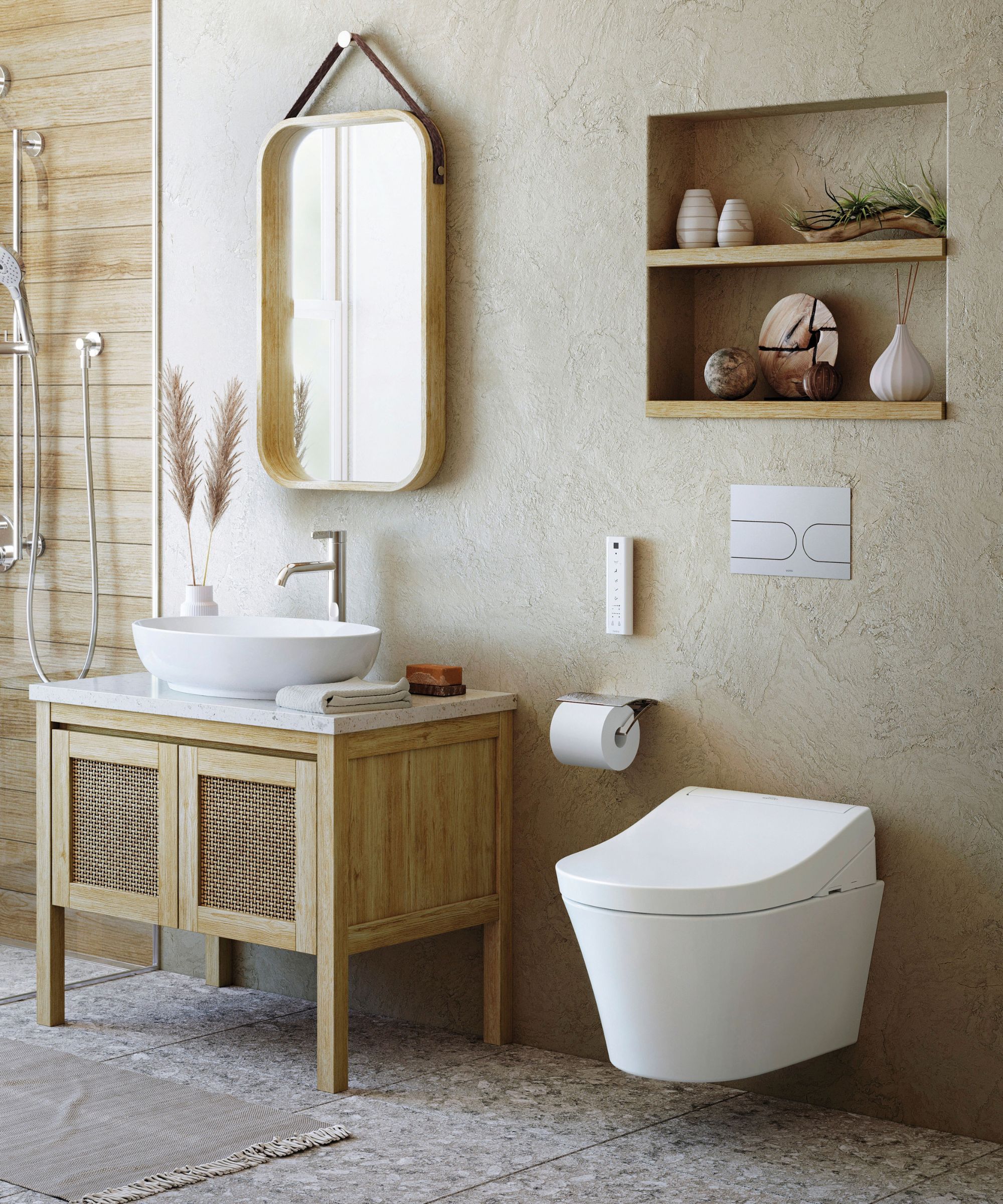 A warm, natural bathroom with a textured sandy plaster wall, a light oak and rattan vanity unit topped with a white vessel basin, and a rounded wood-framed mirror hung on leather straps. A wall-hung smart toilet and recessed oak shelving with decorative objects sit alongside.