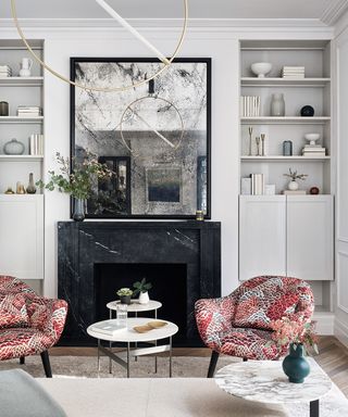 A faded mirror over a black fireplace in a white scheme with red patterned armchairs, illustrating small apartment living room ideas.