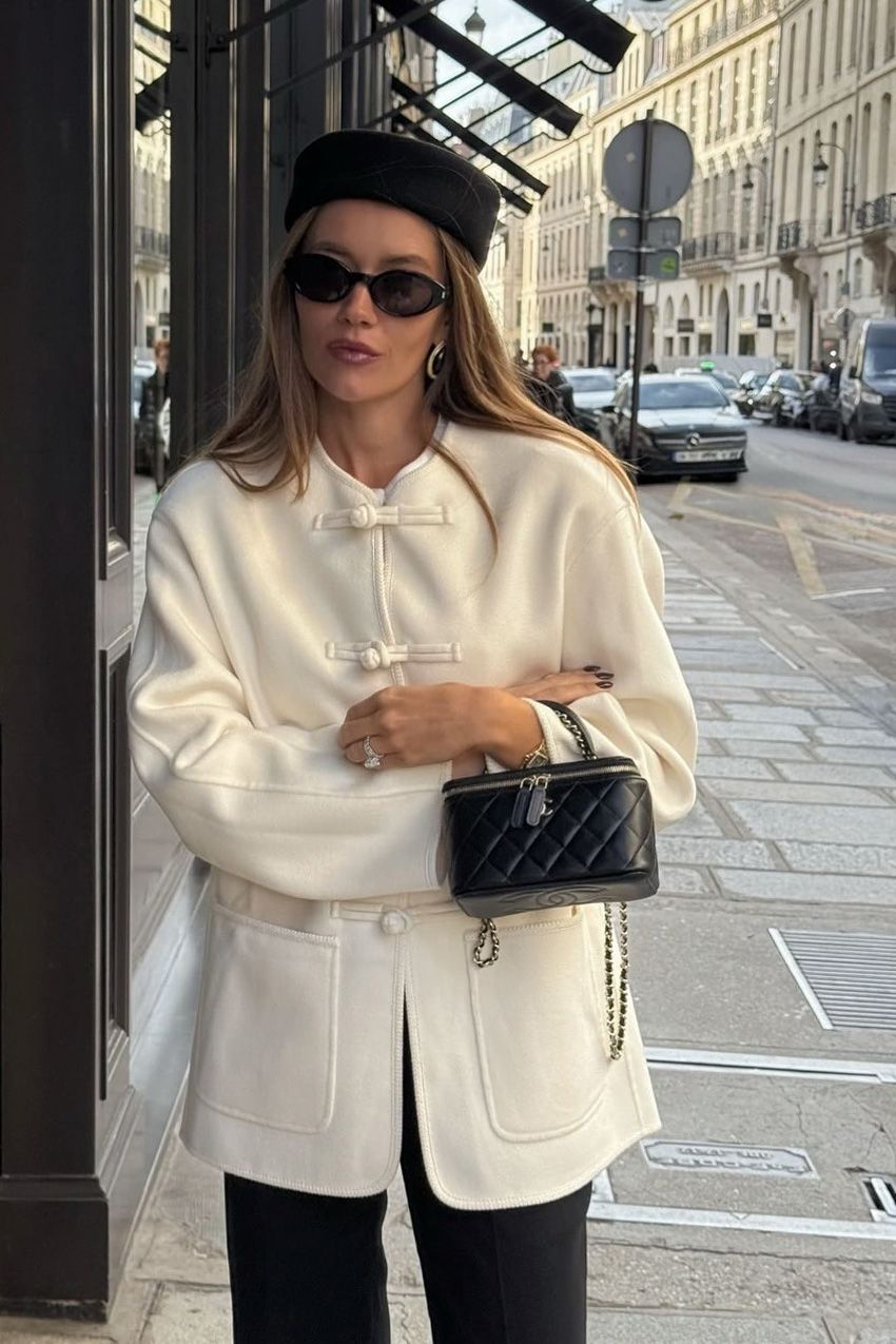 A woman standing outside wearing a white tang jacket styled with black trousers, a black pillbox hat, black sunglasses, and a black Chanel bag.