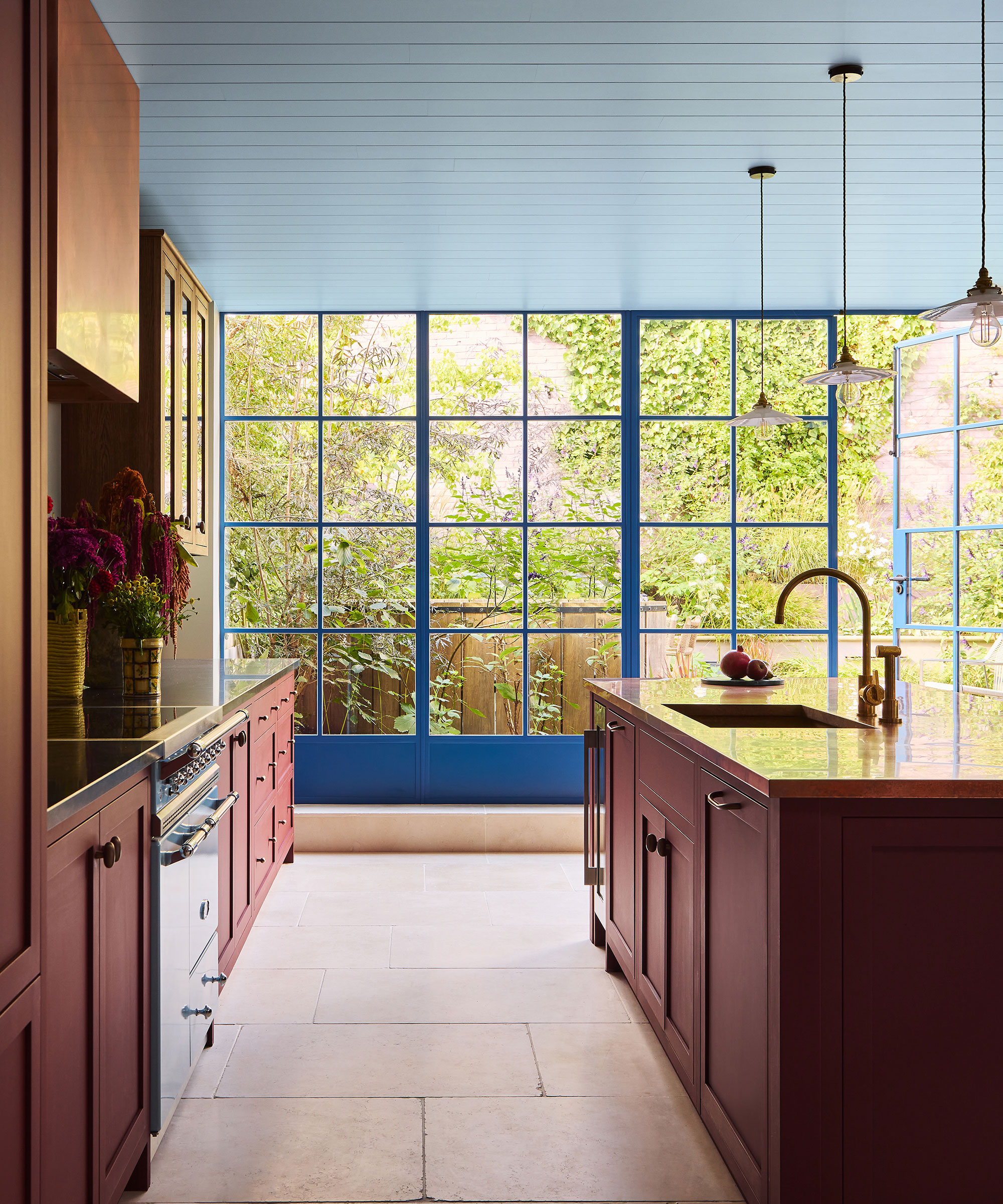 This light-filled kitchen by Tom Morris features striking blue-framed Crittall windows that open to a lush garden, perfectly complementing the sky-blue paneled ceiling. The design is anchored by elegant deep red cabinetry and a large central island, all set against a neutral stone floor for a sophisticated, airy feel.