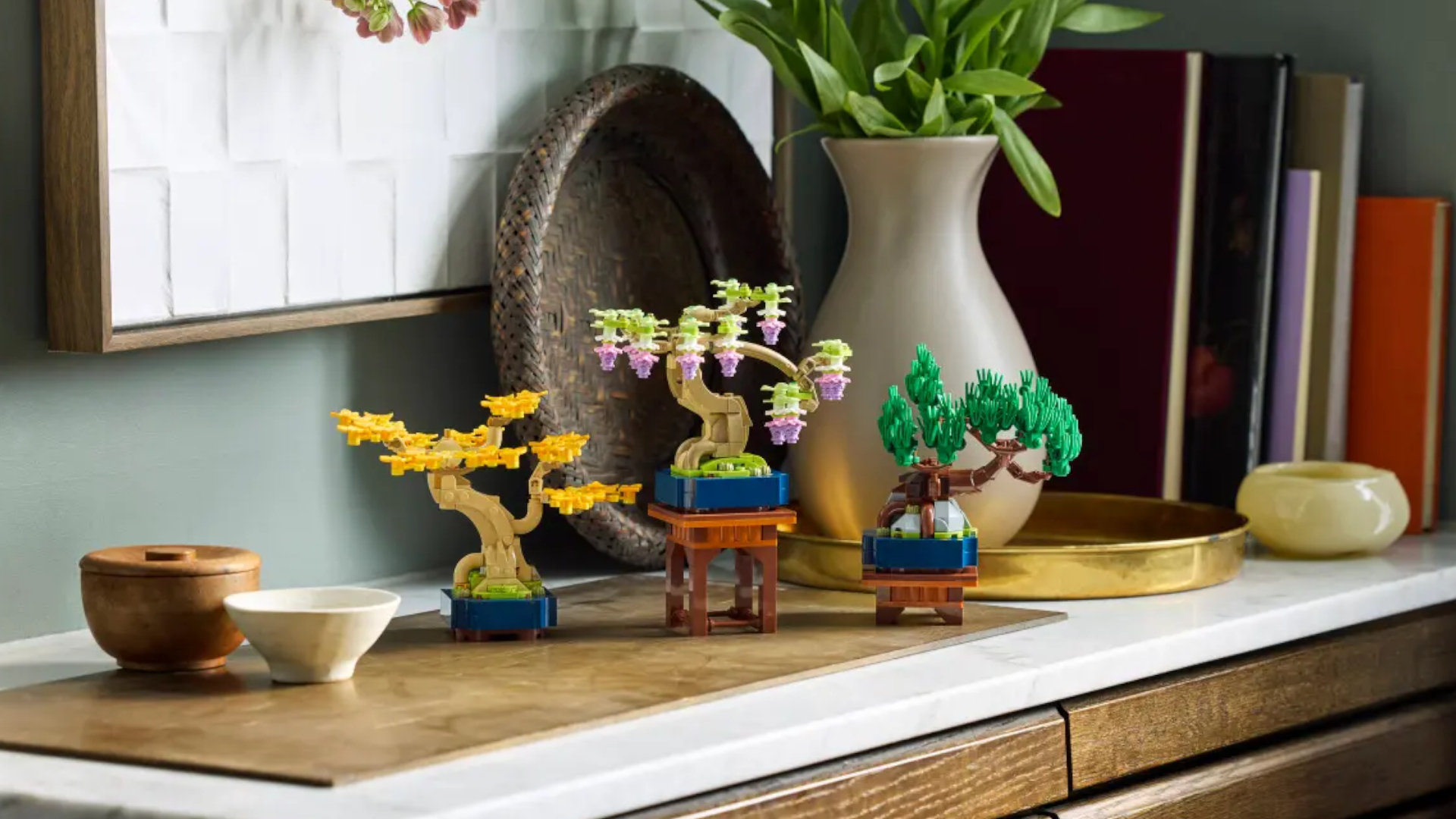 Three miniature Lego trees in pots on a wooden cabinet with a white top, beside bowls, a plant in a vase, and books