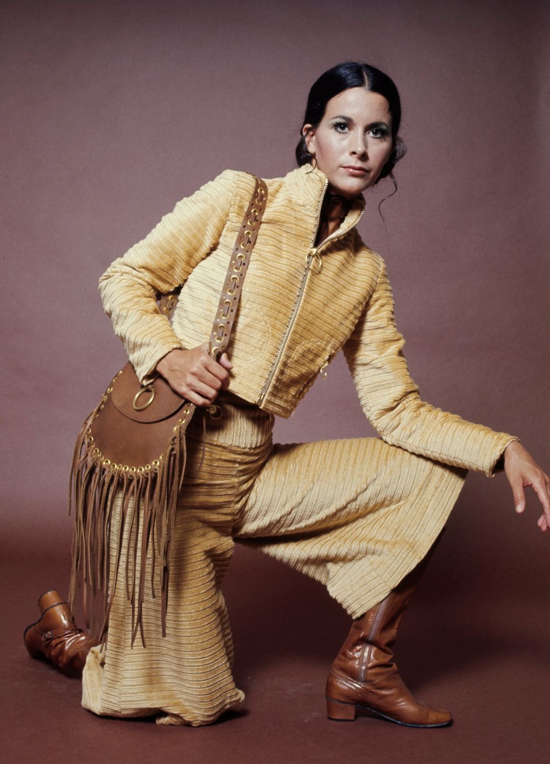 A fashion model wearing a gold-coloured trouser suit with wide-leg pedal pushers and brown boots, with a fringed brown purse, during a studio fashion shoot, against a brown background, United States, circa 1975.
