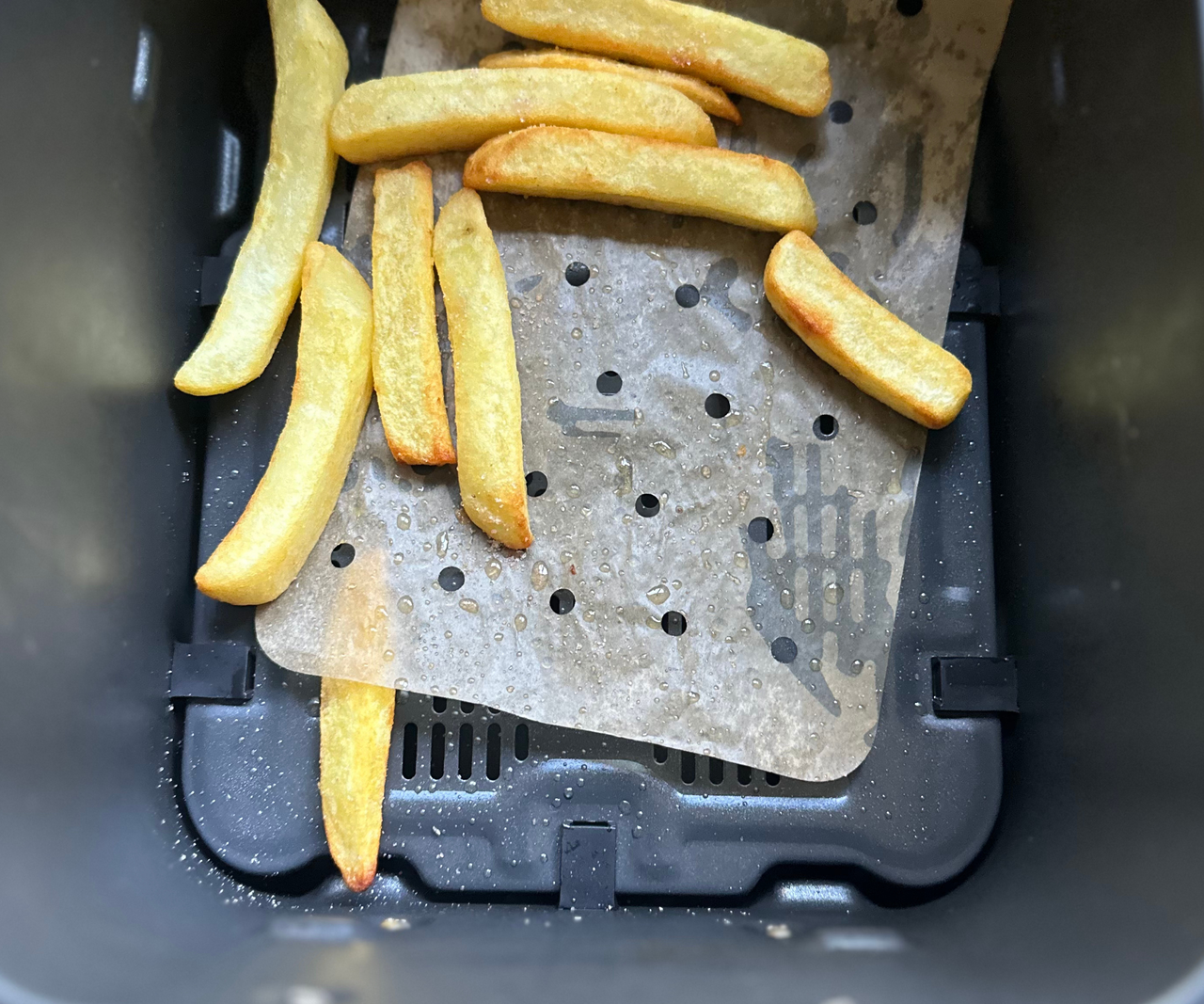 A handful of thick fries on top of a perforated paper air fryer liner inside a black air fryer basket. The liner has shifted so that it sits off-center, and one fry is beneath the edge of it.