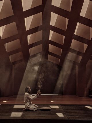 A wellness room with terracotta-hued ceilings and floors is captured as beams of light hit its ground via square top windows while a man dressed in white with his legs crossed holds a smoking incense bowl in his hands.