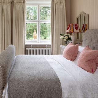 bedroom with grey upholstered bed, grey throw, white bedding, pink cushions, fabric lampshade, cream curtains, cream textured wallpaper