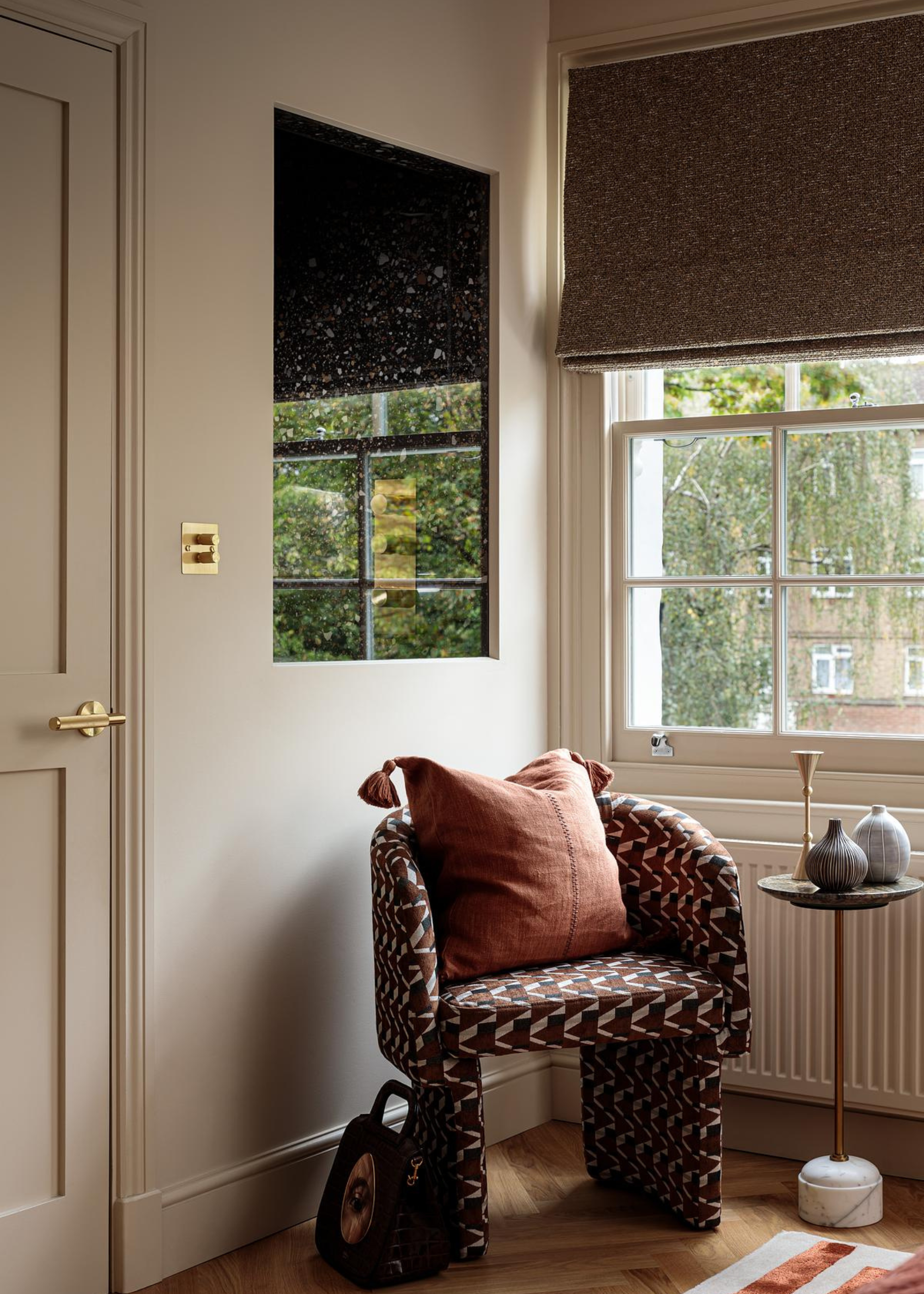 A patterned sofa stool with a tassel pillow near a side table in a window-clad corner