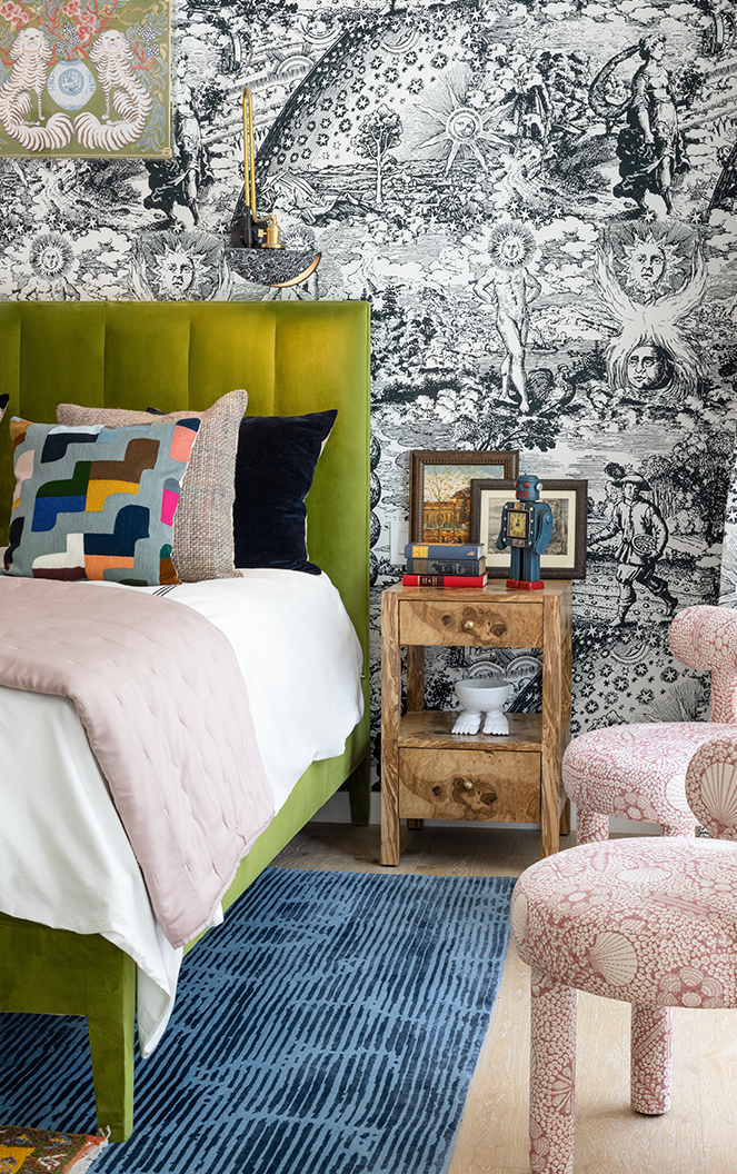 maximalist bedroom with black and white patterned wallpaper, green velvet bedframe, white sheets with colorful throw pillows, timber nightstand, blue striped rug under the bed, two upholstered pink patterned armchairs, and lots of picture frames on the bedside table