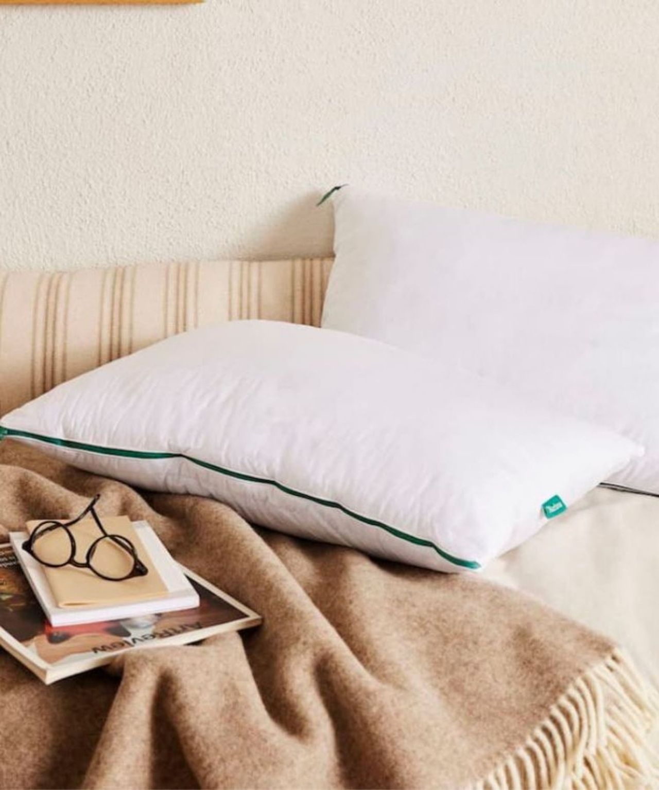 Two Brooklinen Marlow Pillows pictured in a neutral bedroom on a bed with striped headboard, with neutral bedding, a warm brown blanket, and a stack of magazines and notebooks with black glasses.