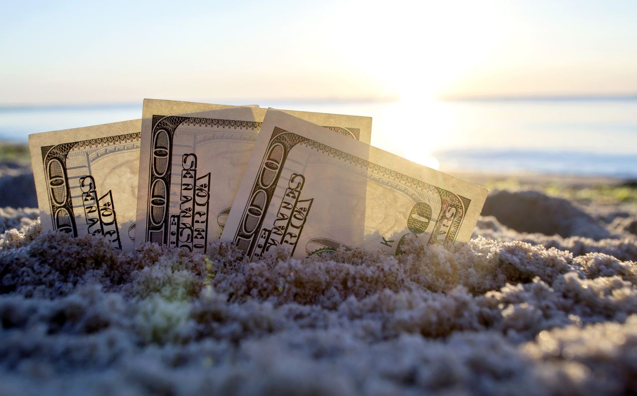 three one hundred dollar bills in the sand with a shoreline in the background