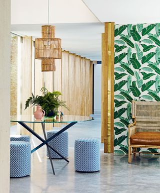 Three blue and white stools with geometric pattern around circular glass table on screed floor, rattan pendant lamp and rattan sofa, banana leaf wallpaper.