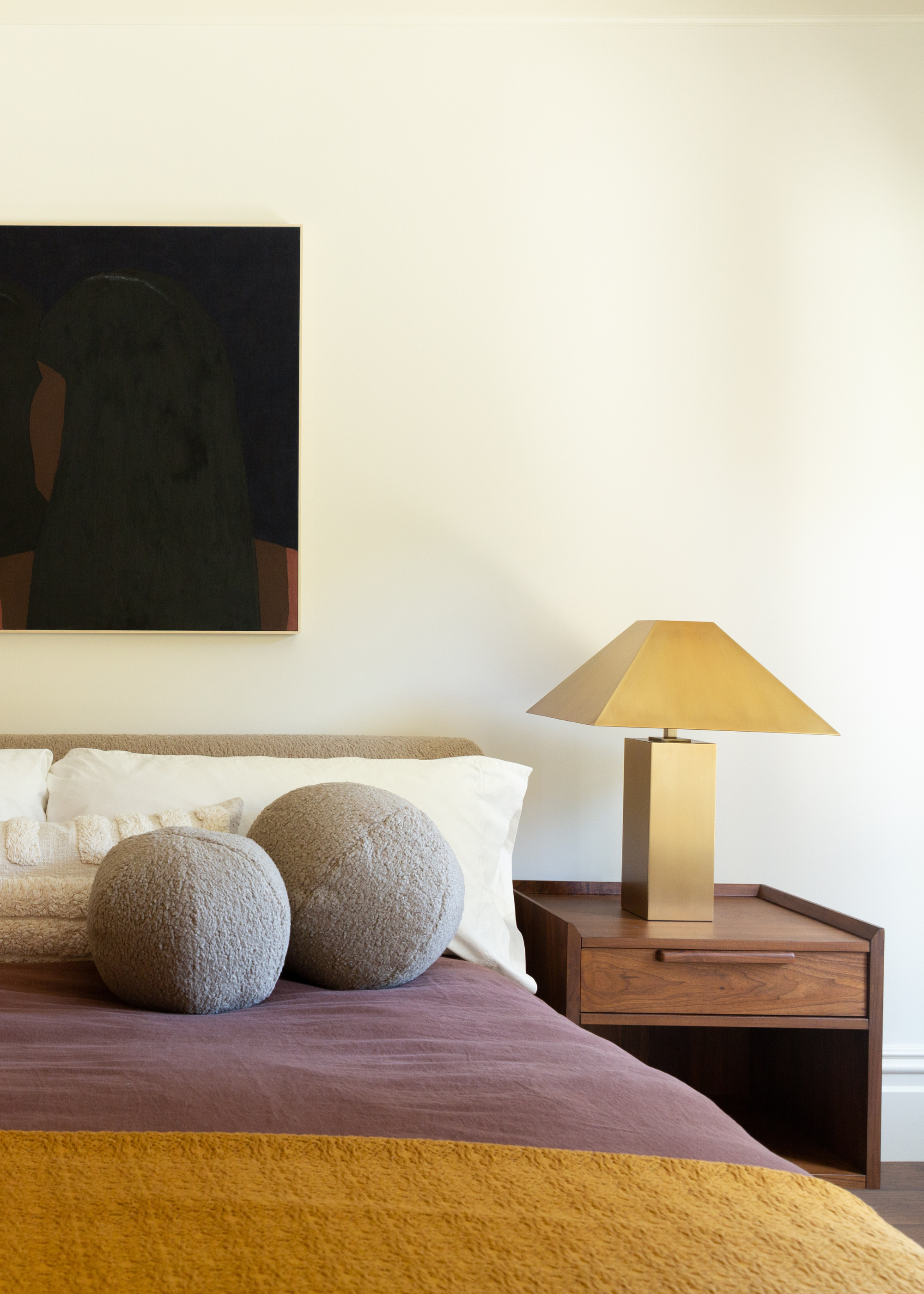 A white bedroom with art on the wall, a wood nightstand with a gold lamp and a bed with gray round pillows on a purple duvet