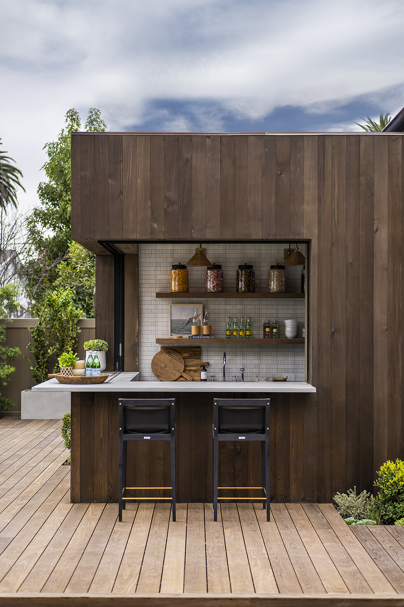 an outdoor kitchen covered by a wooden structure with stone counters and a tiled wall behind