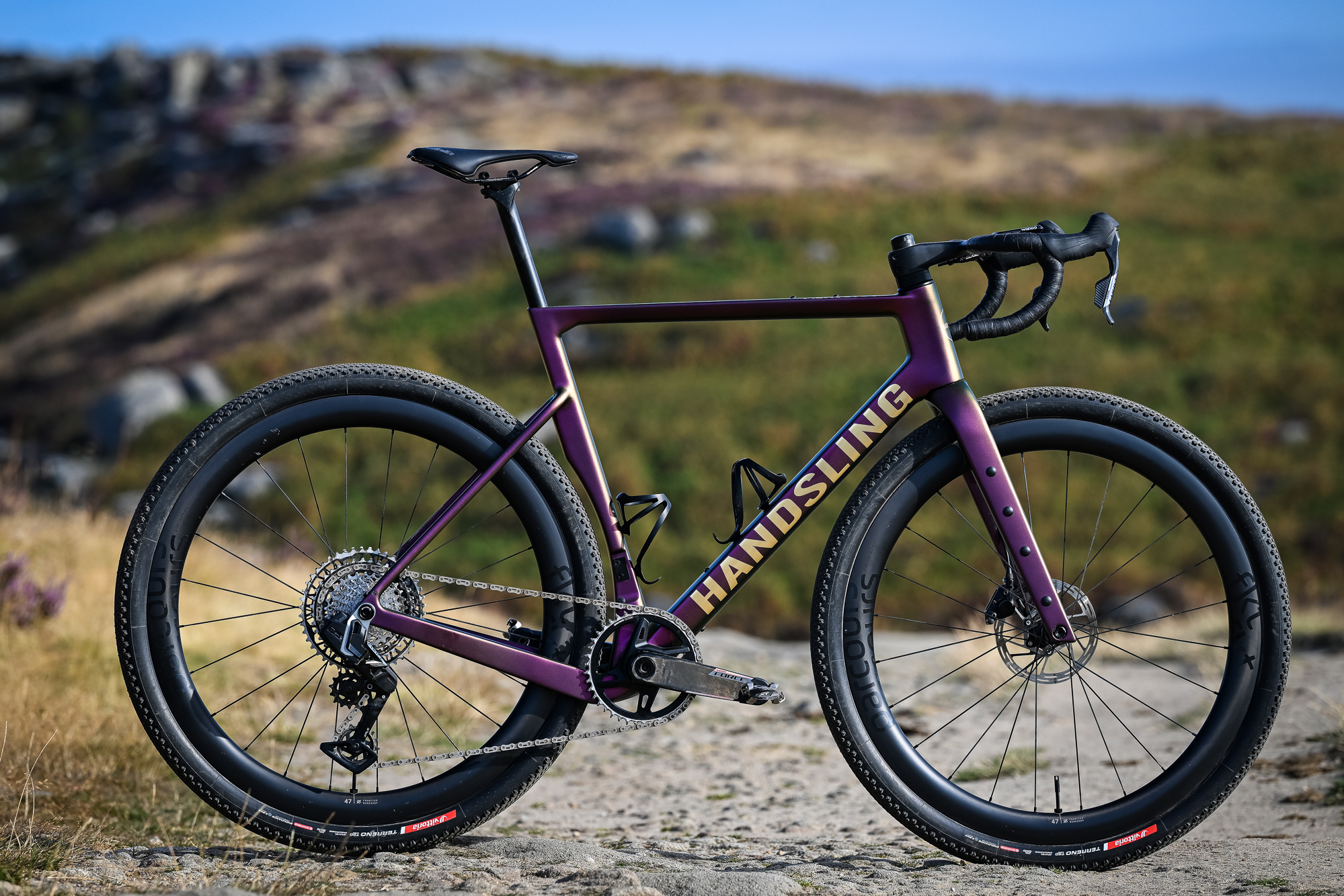 A purple gravel bike side-on to the camera on a sandy trail with a crag in the background and 'Handsling' written on the downtube.