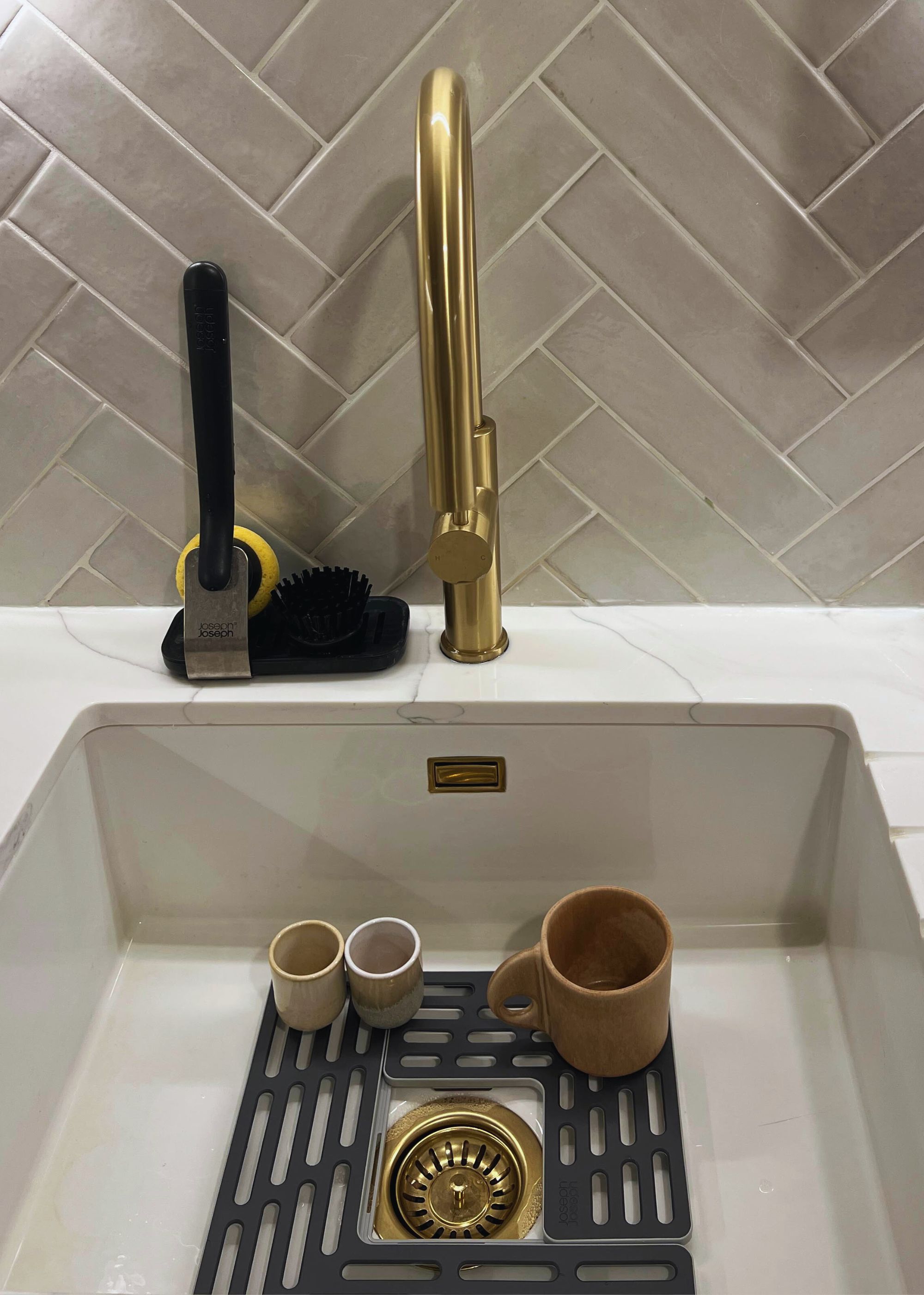A sink protector placed inside a sink with a mug and espresso shot glasses inside. There is also a brass gold kitchen tap with a sponge on the left side of the tap.