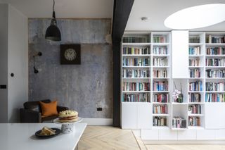 Wall of rear extension with built-in bookshelves under round skylight, and small reading look with concrete-effect wall, black pendant light, black wall-hung reading light and brown leather armchair