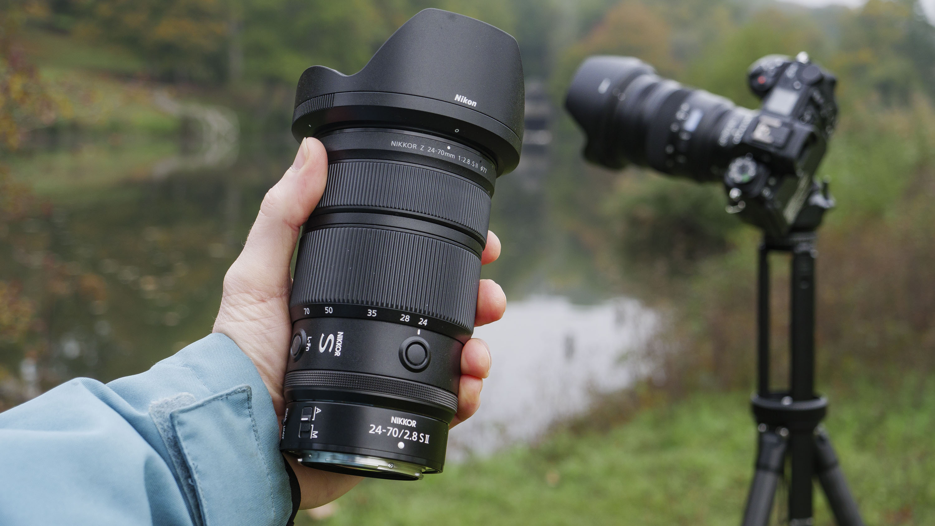 Nikon Z 24-70mm f/2.8 S II lens in user&#039;s hand, with another Nikon lens mounted to a tripod with autumnal lake background