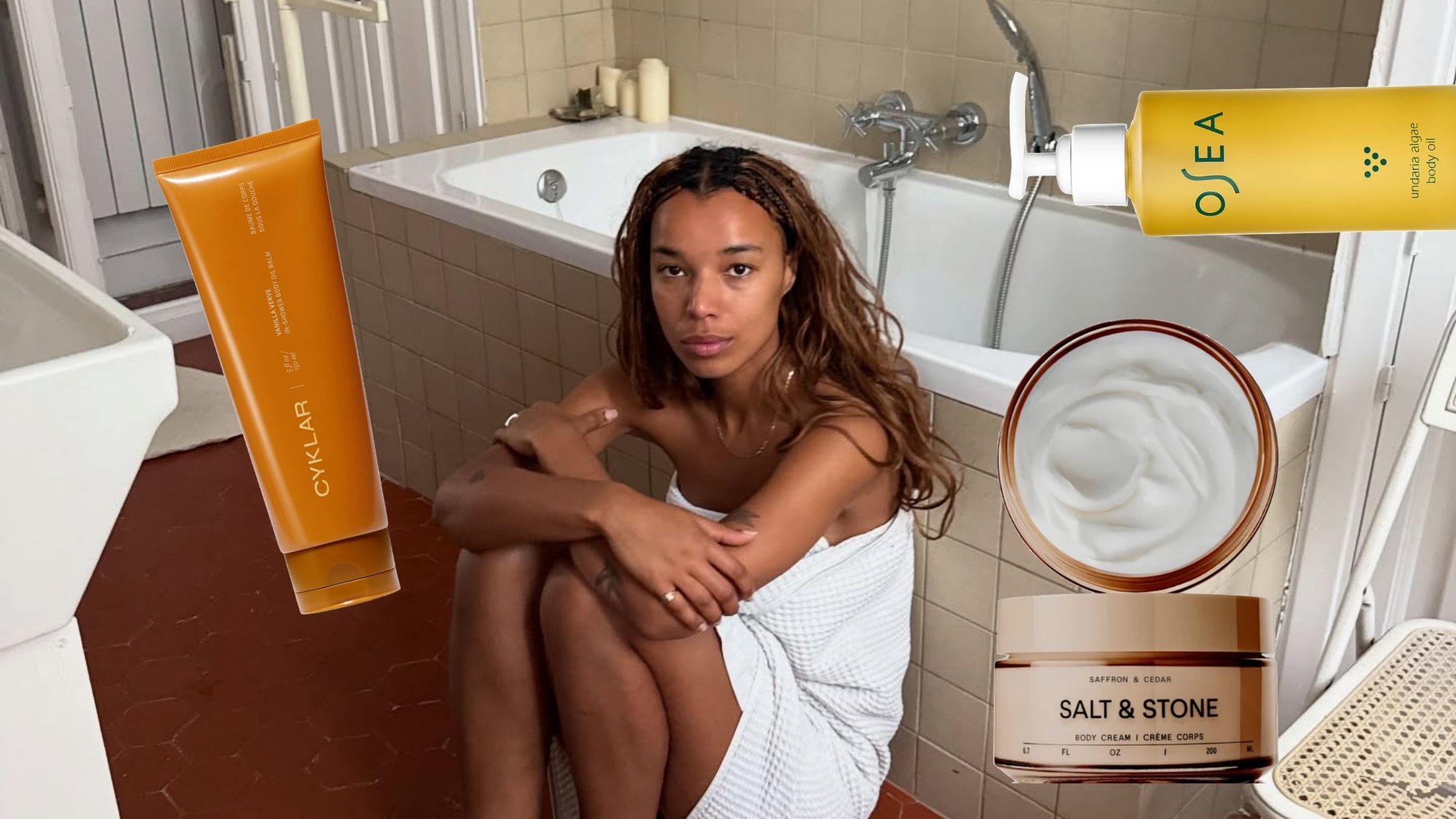 A woman in a towel leaning against a bathtub surrounded by products.