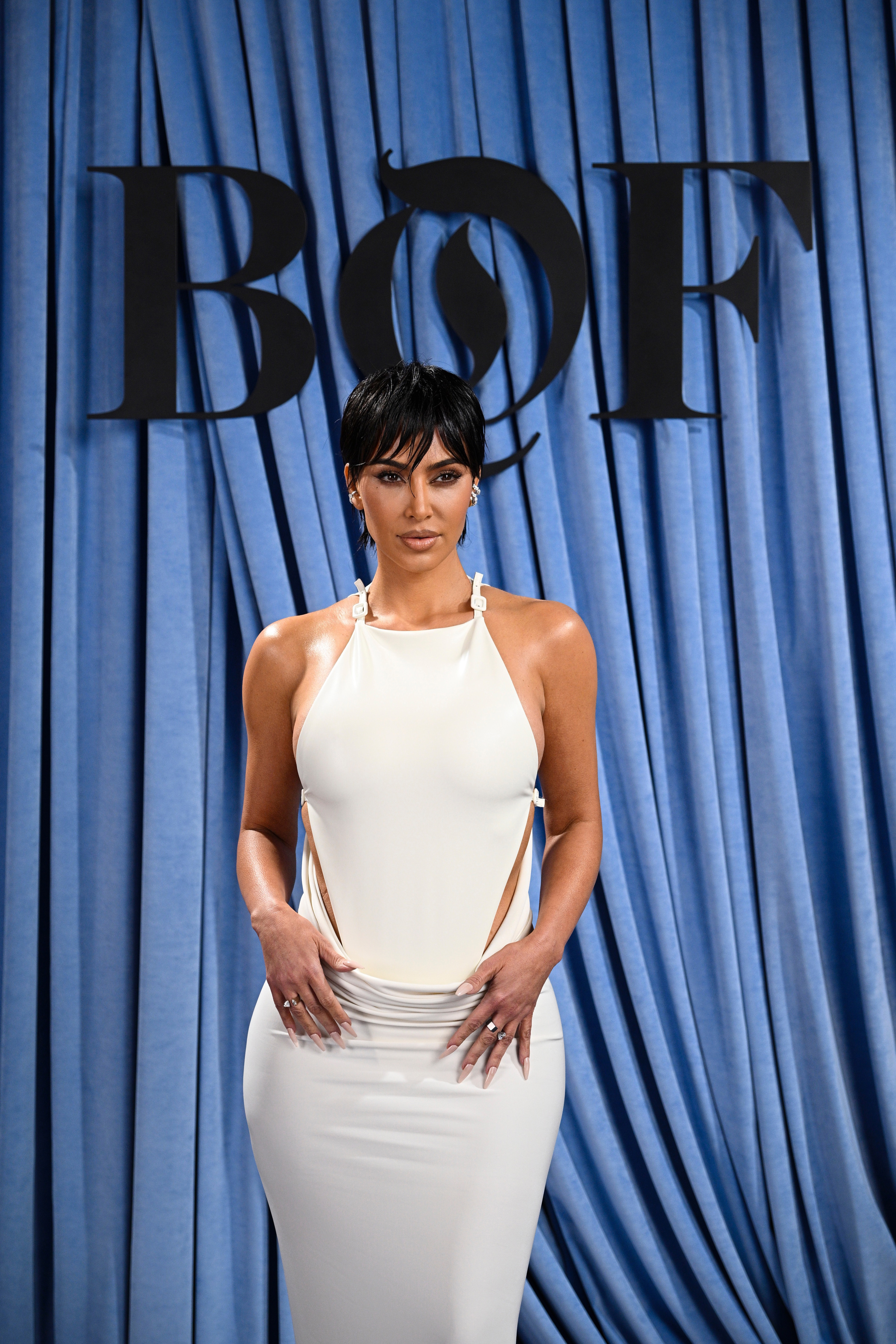 a woman with a short haircut wearing a white halter dress in front of a blue curtain