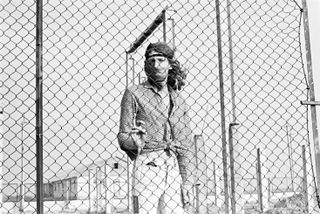Man with long hair standing behind a high, wire fence