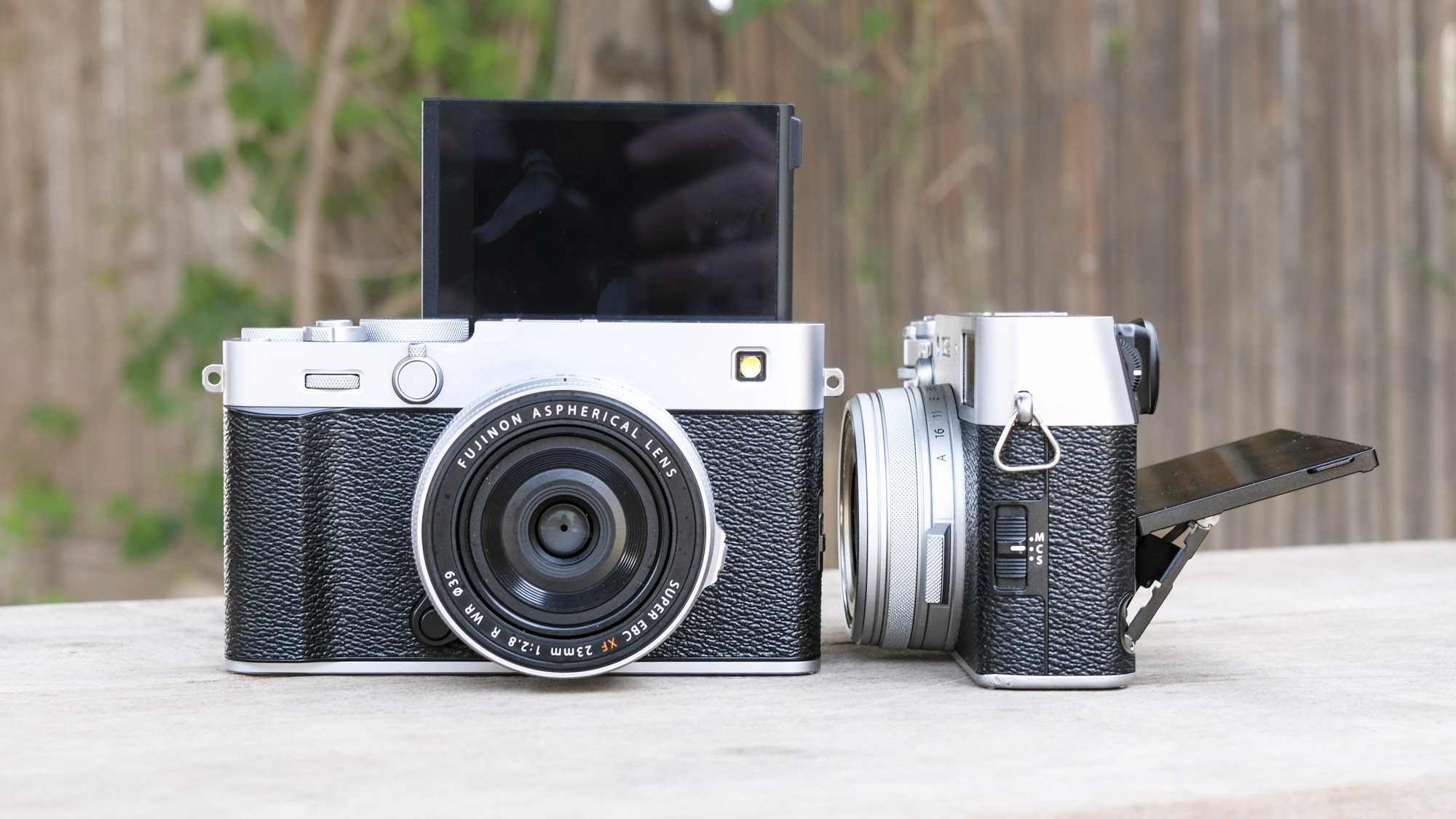 Fujifilm X-E5 with its screen in selfie mode, alongside the Fujifilm X100VI with screen flipped up, on a wooden bench