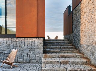 Close up view of the stone steps and Corten steel and stone exterior at Smith House. There is a wooden chair on the lower level and two wooden chairs on the upper level