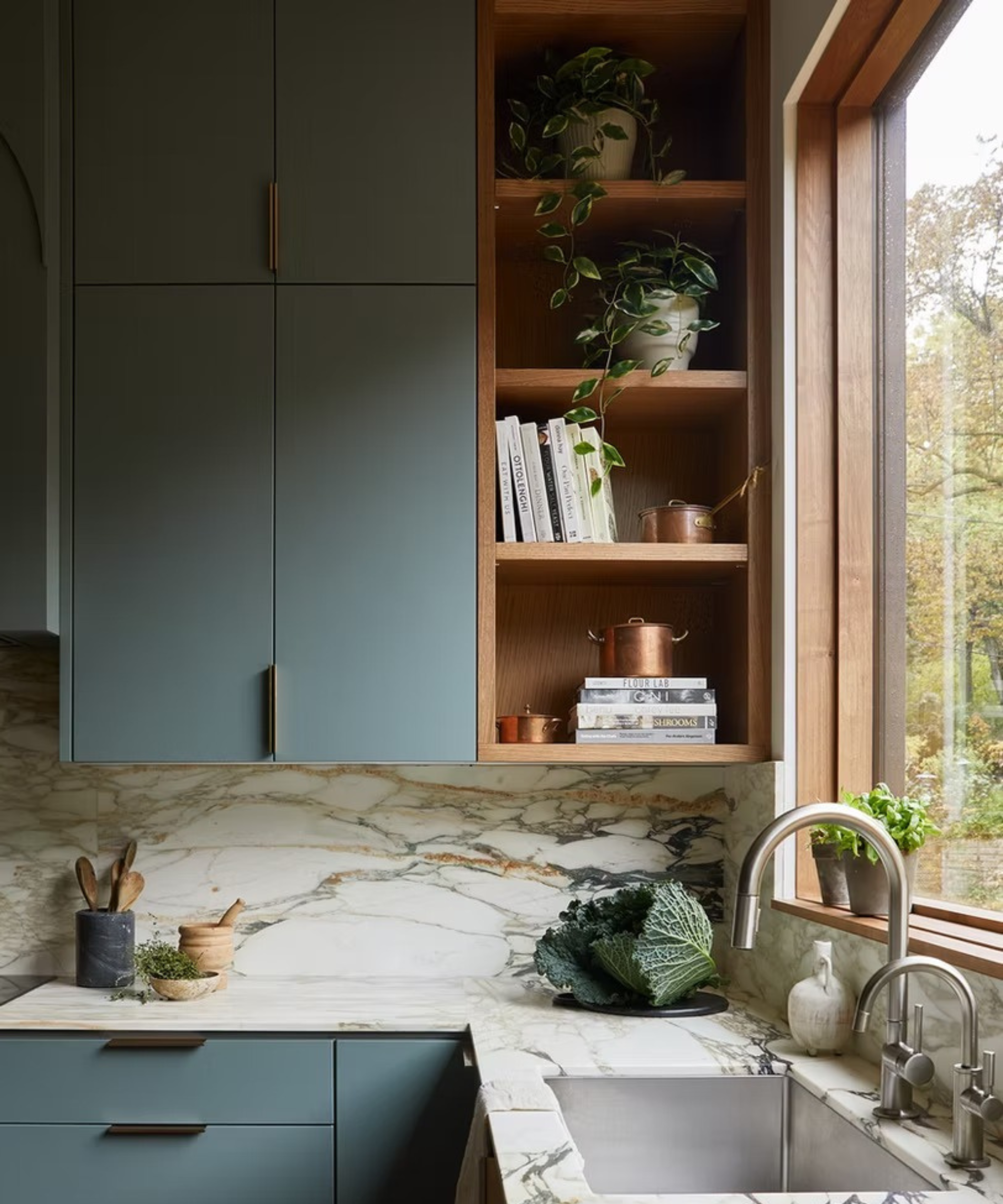 a kitchen with white and gray marble counters and backsplash, grayish blue painted cabinets, an open shelving unit with books and plants, and cabbage on the countertop