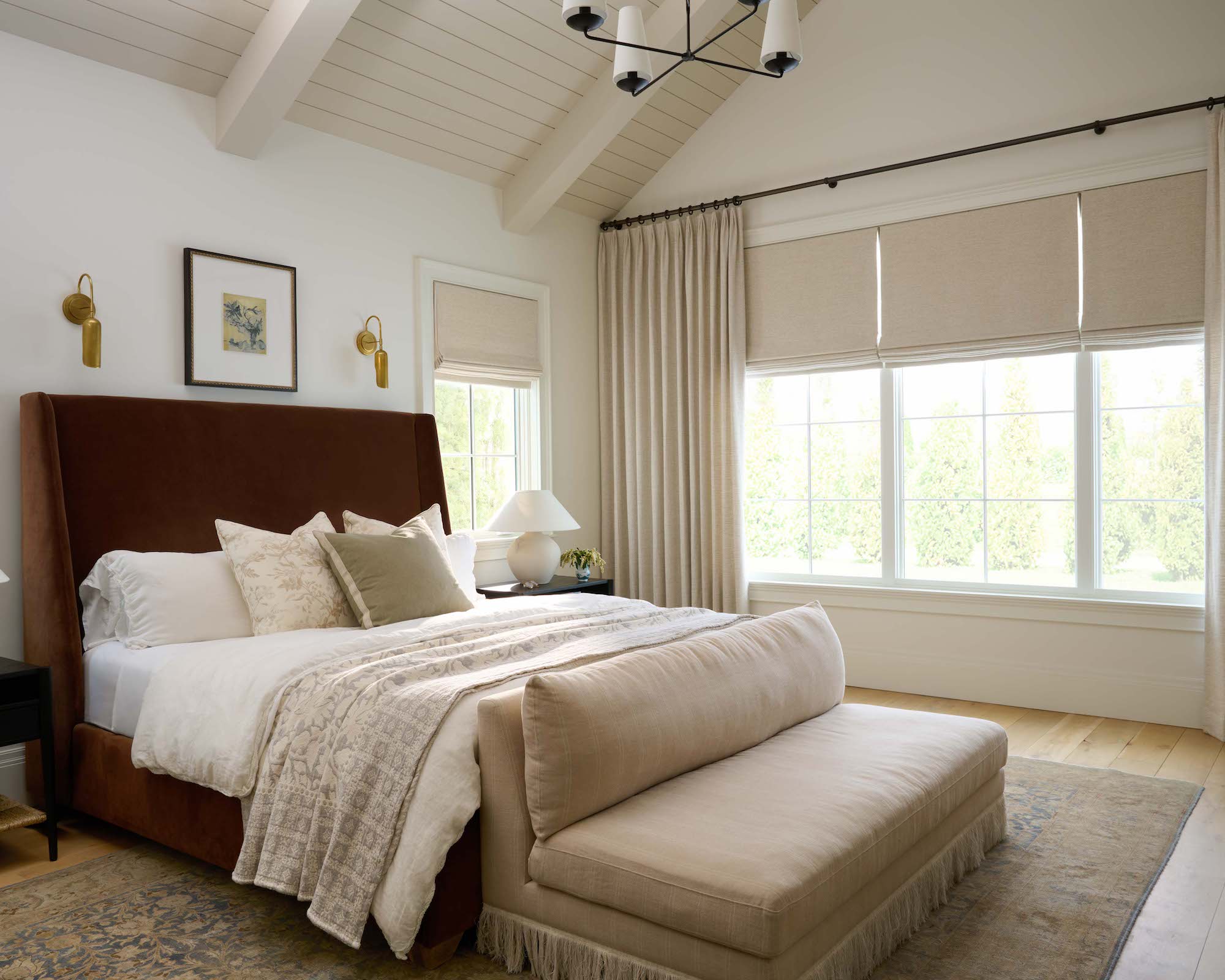 Neutral bedroom with sloping ceiling and wood floor with area rug, bed dressed in neutral linen, couch, and windows dressed with Roman blinds and curtains