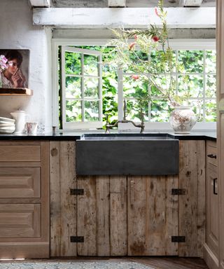 Rustic sink area with large stone farmhouse sink, open window onto greenery, and rustic reclaimed wood cabinets.