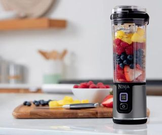 A press image of the Ninja blast filled with fruit in a modern kitchen. Behind it is a wooden chopping board with cut fruit