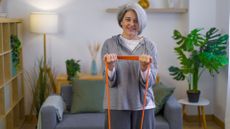 woman facing the camera dressed in grey slouchy clothes curling an orange resistance band toward her chest. there's a grew sofa and a living room setting behind her.