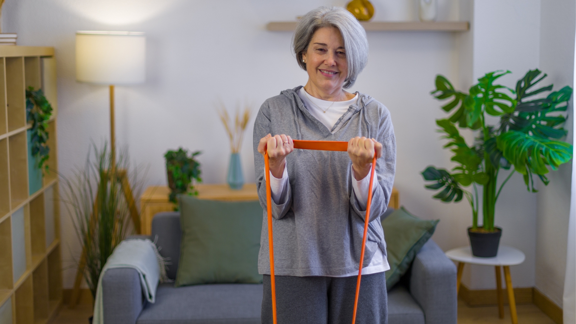 woman facing the camera dressed in grey slouchy clothes curling an orange resistance band toward her chest. there's a grew sofa and a living room setting behind her.