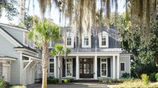 Colonial-style home with Boothbay Gray painted exterior, large porch with dark brown front door, a white garage to the left, and a weeping willow and palm tree at the front of the home