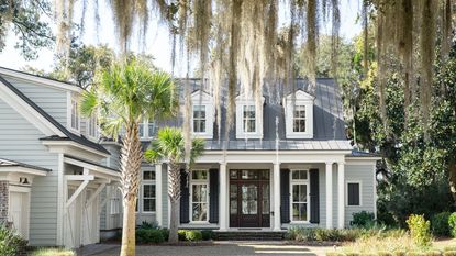Colonial-style home with Boothbay Gray painted exterior, large porch with dark brown front door, a white garage to the left, and a weeping willow and palm tree at the front of the home