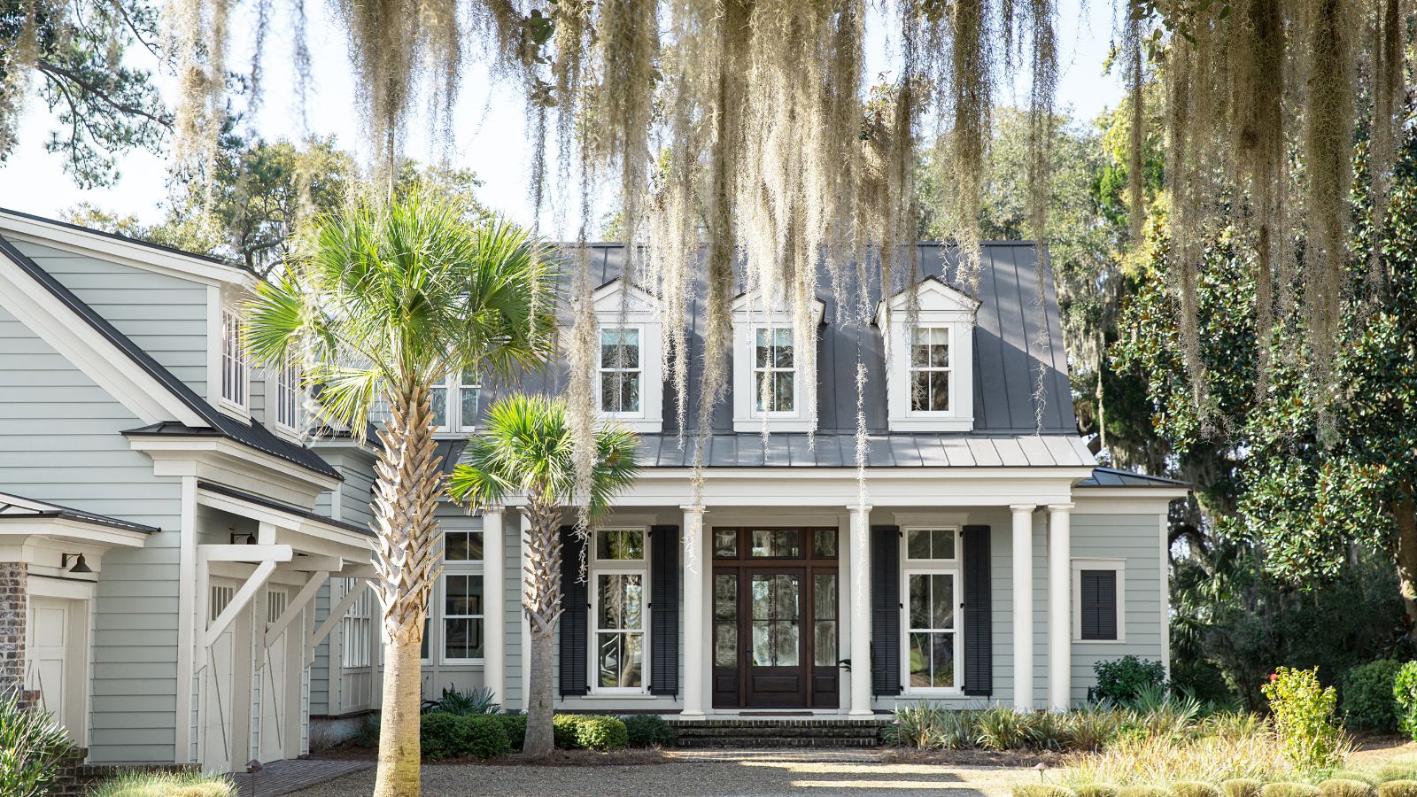 Colonial-style home with Boothbay Gray painted exterior, large porch with dark brown front door, a white garage to the left, and a weeping willow and palm tree at the front of the home