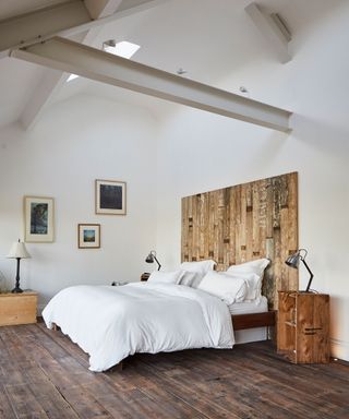 Large room in converted barn, dark wood reclaimed flooring, white painted walls and white painted metal beams, wooden headboard and bedside table, three framed images on wall, table lamps dotted around the room