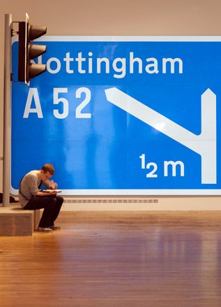 Visitor sitting on wooden stage in front of robot and blue and white road sign to Nottingham