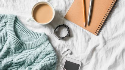 A white bed with a blue sweatr, cup of coffee, wate, and notepad with pens laid on it, close up