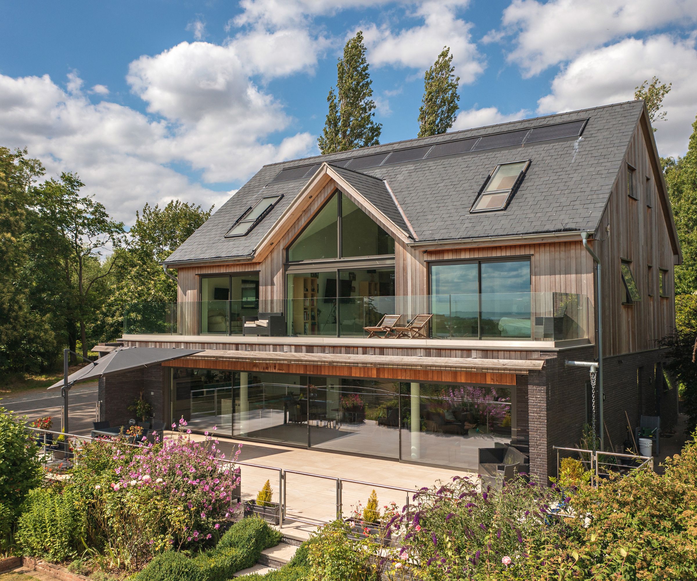 Modern timber-clad house with large glass balcony, slate roof with solar panels, and lush landscaped gardens bursting with flowers.
