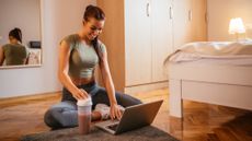 Woman sitting on the floor in front of a laptop and protein shaker filled with a red-purple liquid