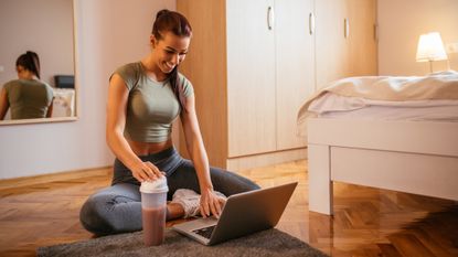 Woman sitting on the floor in front of a laptop and protein shaker filled with a red-purple liquid