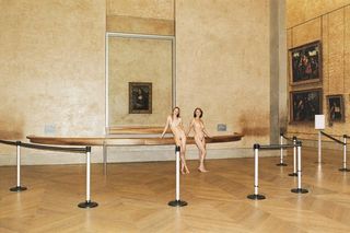 Two slim ladies pose naked in an art gallery with high ceilings and a wooden floor.