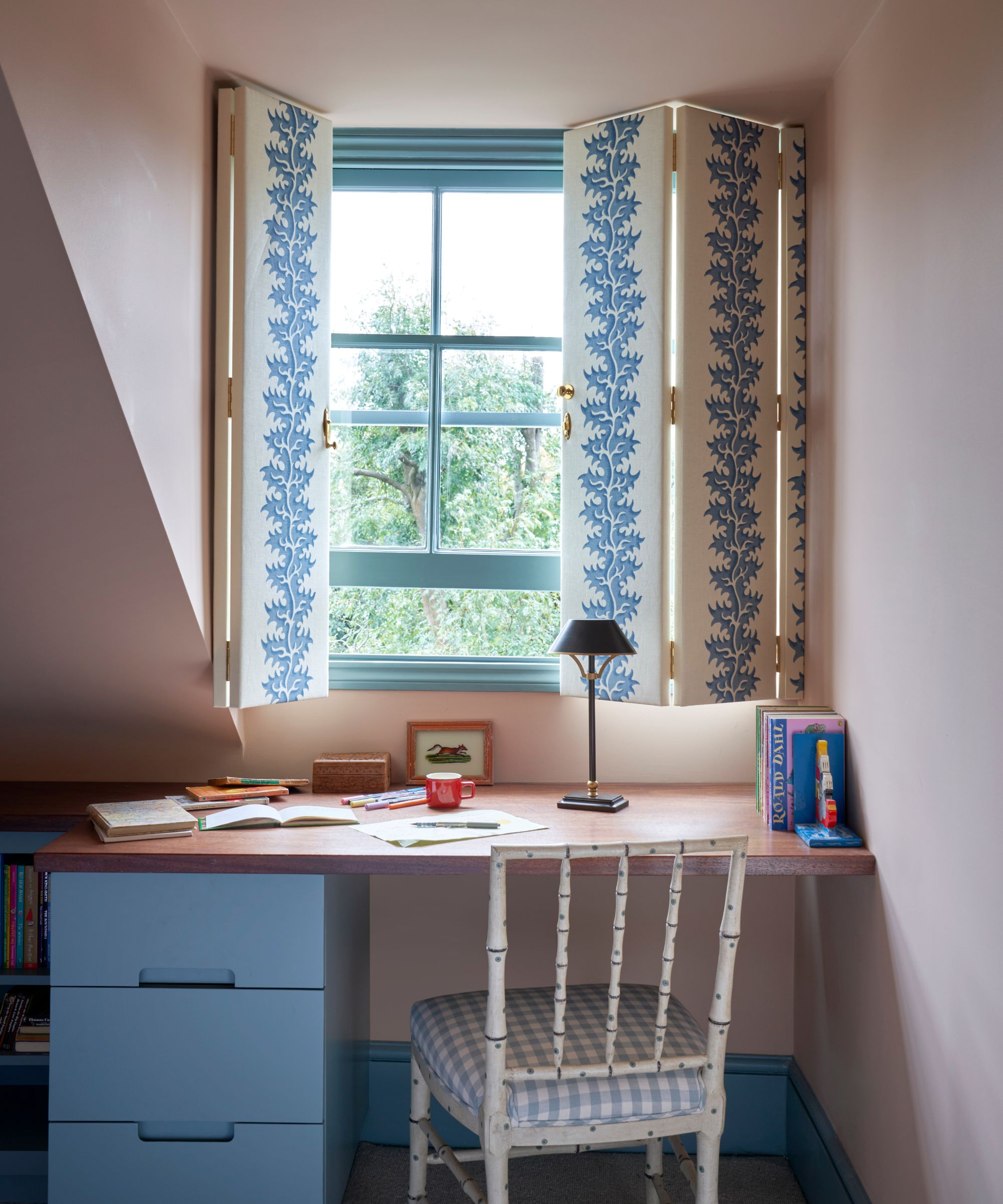 Home office with pale pink walls, pale blue painted window with fabric folding window blind, wooden desk with blue drawers, and a white-painted bamboo chair with a gingham seat