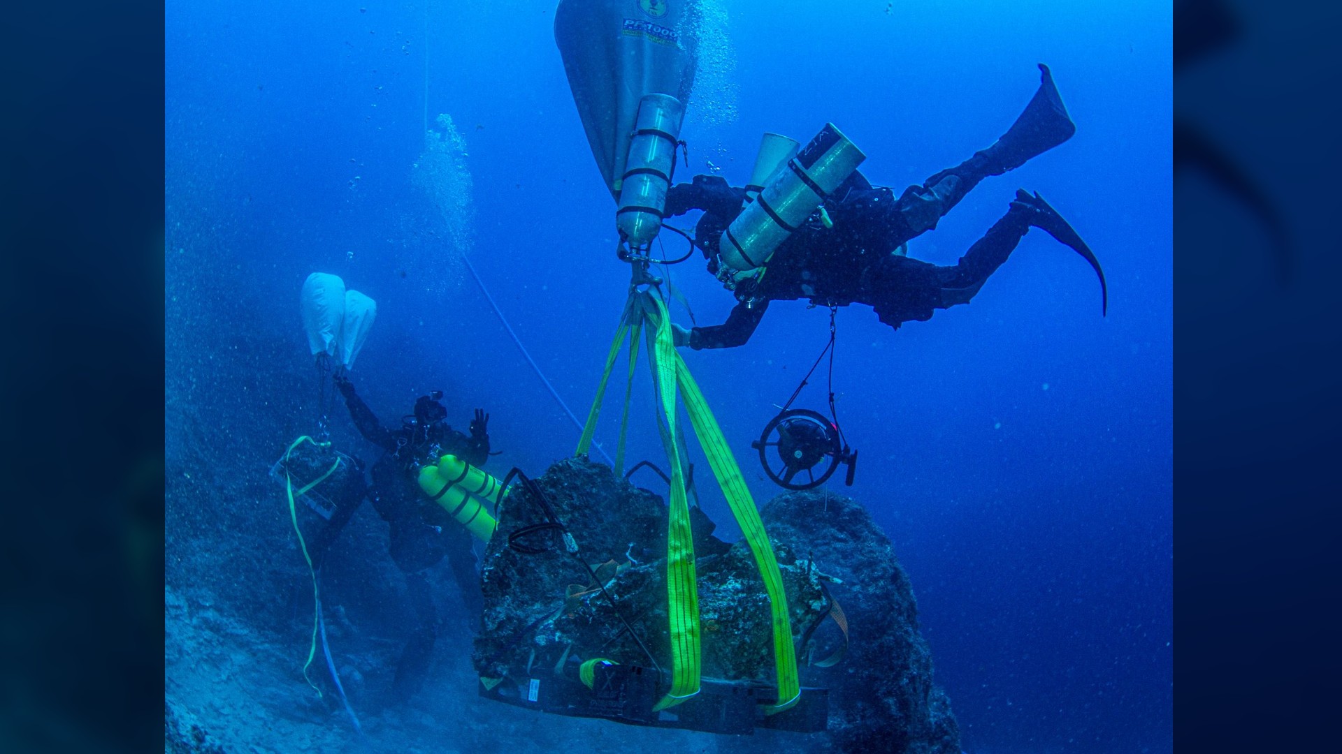 Here we see 2 scuba divers underwater using some kind of balloon and long yellow ropes to wrap around a badly corroded marble plinth that was found at a shipwreck underwater.