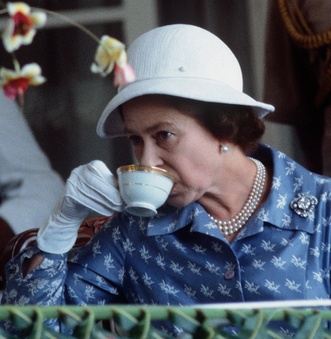Queen Elizabeth wearing a blue dress and white hat drinking tea from a cup