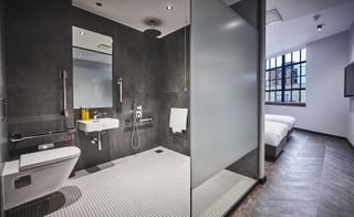 A bathroom in the New Road Hotel. Large gray tiles cover the walls, while small white tiles cover the floor. The show is completely open. A toilet is to the left, and a sink with a large mirror next to it.