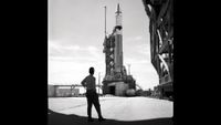A man stands in front of a rocket on the launch pad in black and white.