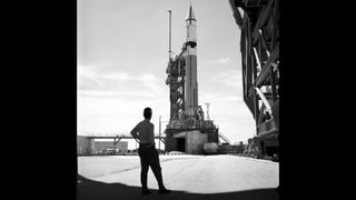 A man stands in front of a rocket on the launch pad in black and white.